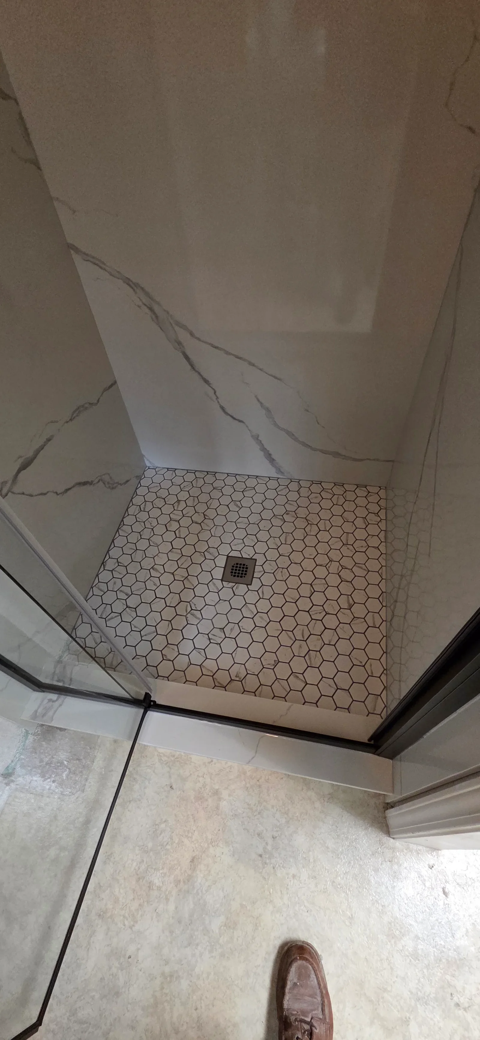Walk-in shower with marble-look wall panels, hexagonal white mosaic floor tile with dark grout, brushed metal drain, and black-framed glass door