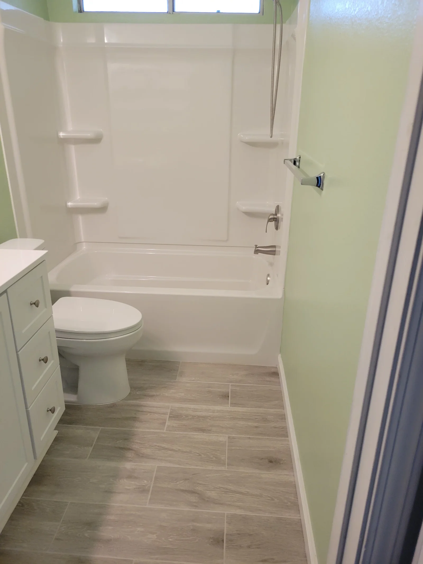 Bathroom with white tub-shower surround, wood-look gray tile floor, sage green walls, and brushed nickel fixtures