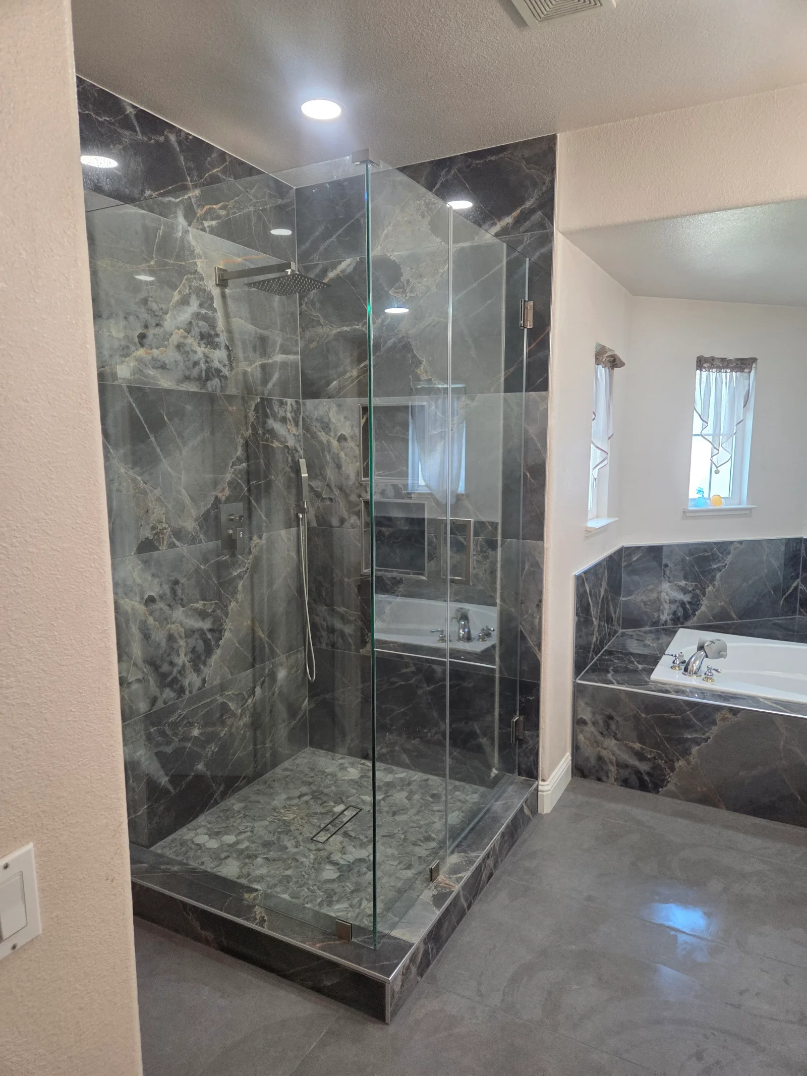 Walk-in shower with frameless glass door, dark gray marble-look large-format tile, hexagon mosaic floor, and tiled drop-in tub surround