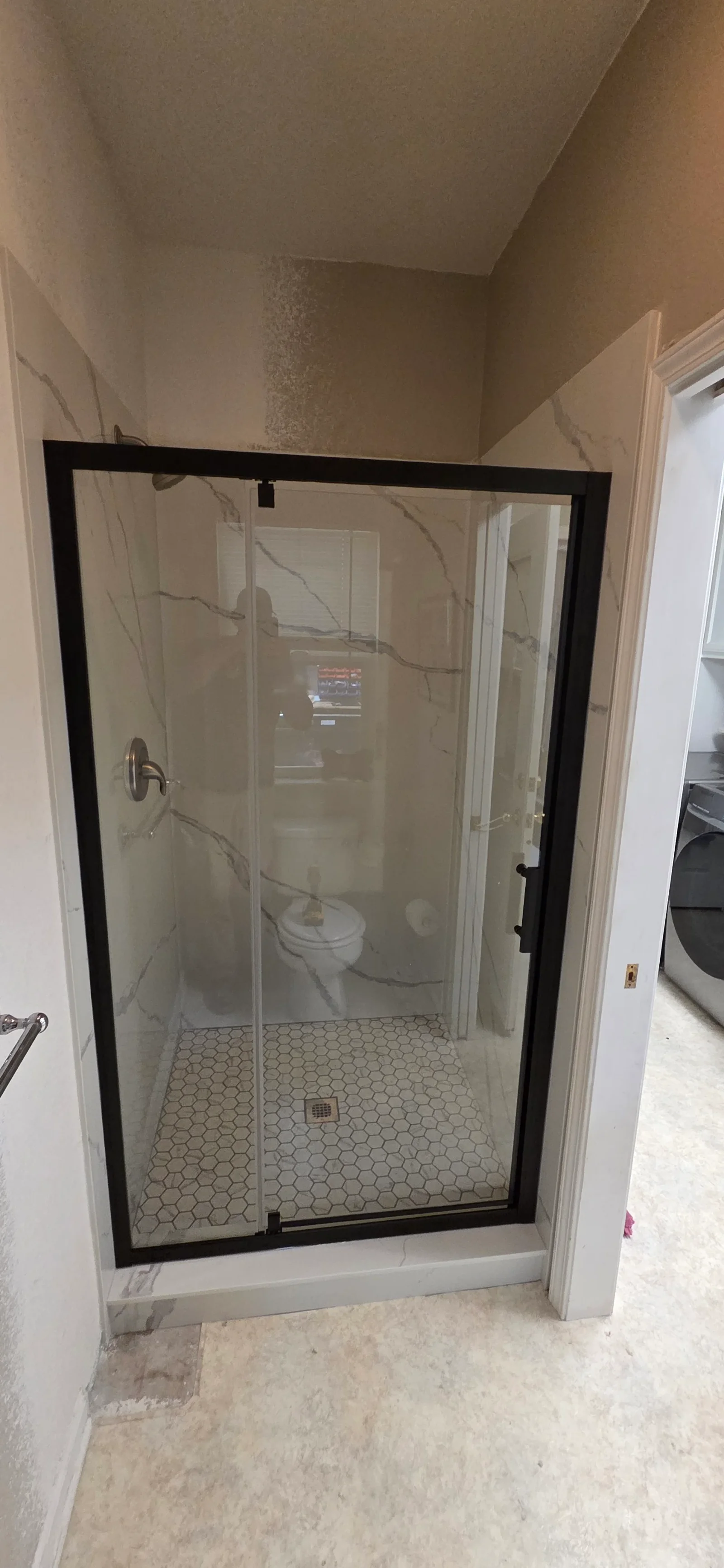 Shower with black-framed glass door, white marble-look wall panels, and white hexagon floor tile with brushed nickel drain