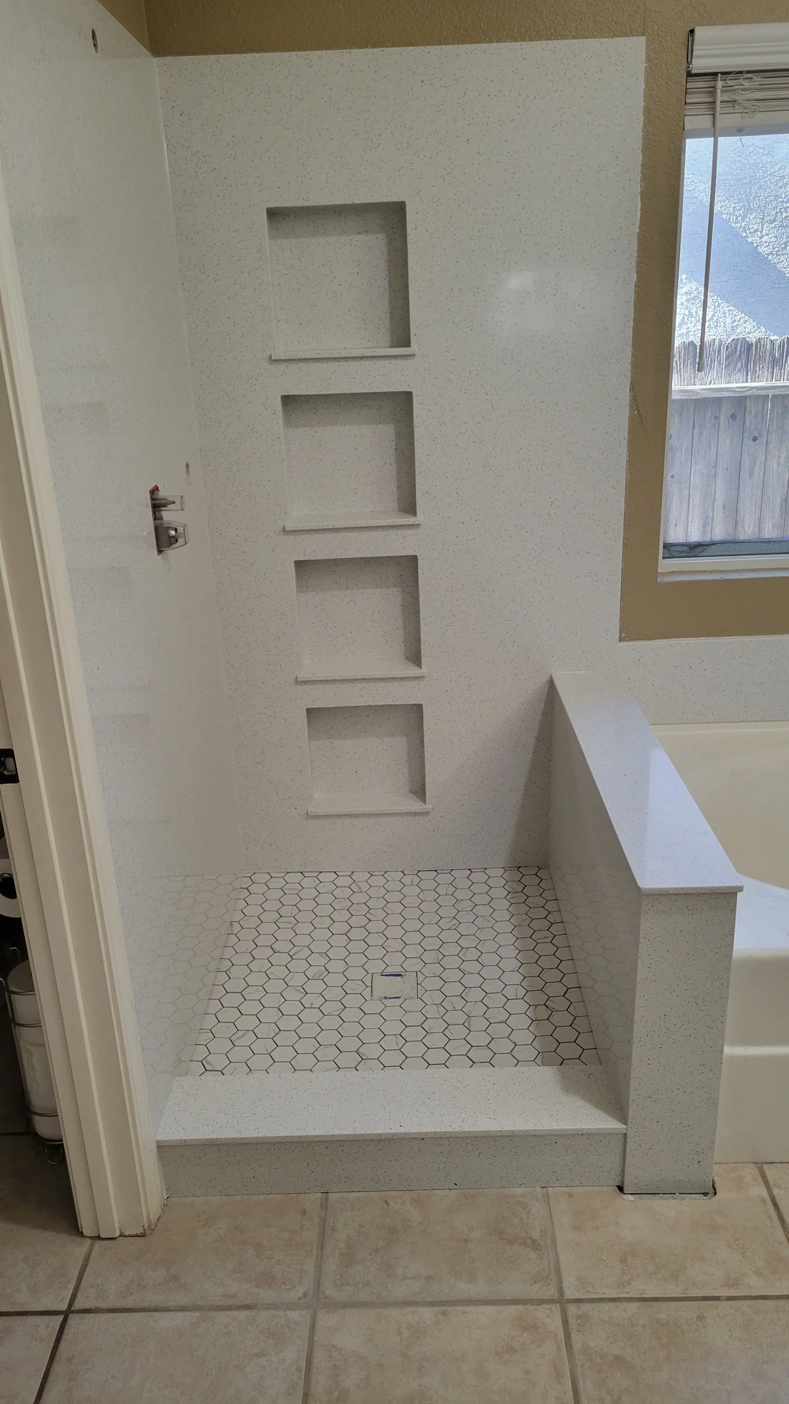 Walk-in shower with speckled white solid-surface walls, four stacked recessed niches, and white hex tile floor with dark grout