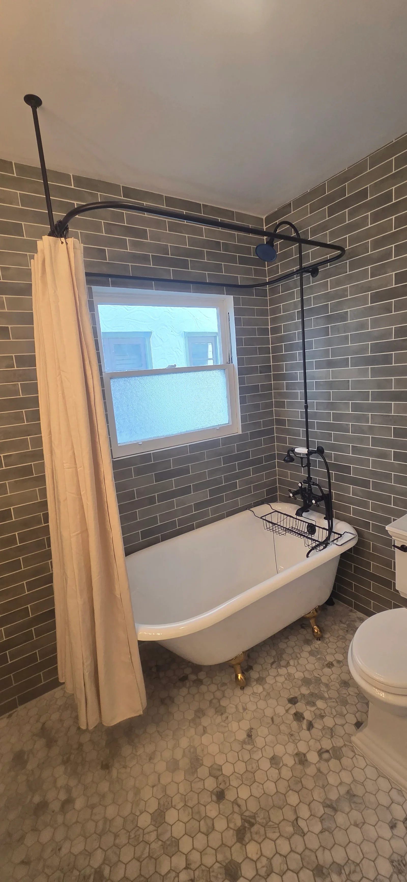 Clawfoot tub with black exposed shower riser, gray subway tile walls, and white hexagon floor tile