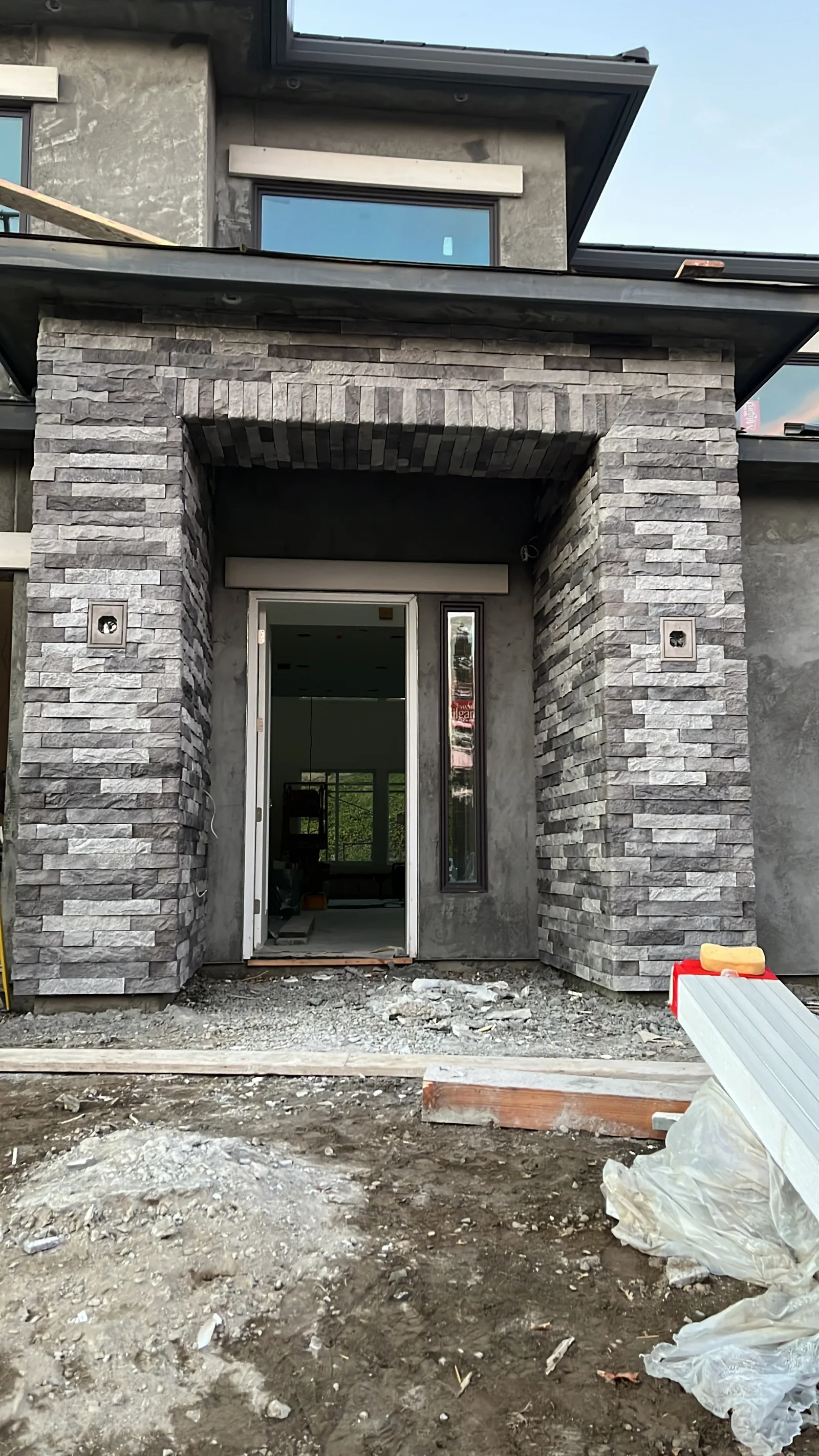 Gray stacked stone veneer entryway surround with arched header, stucco walls, and dark-framed sidelight window during construction
